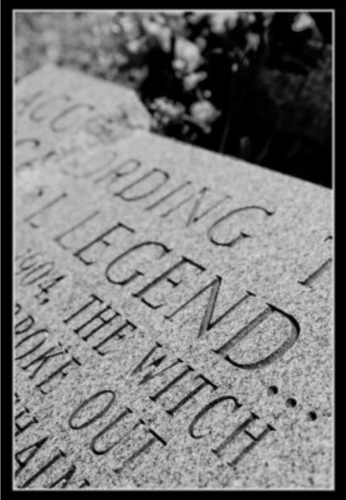 Close-up of a weathered stone with engraved text about a local legend, partially obscured by foliage.