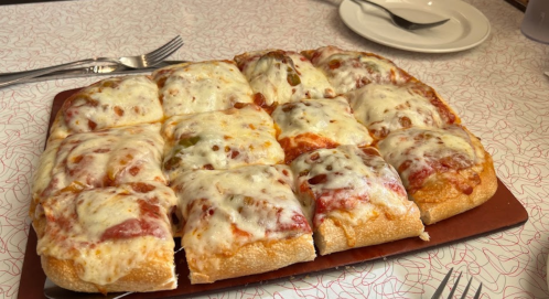 A rectangular pizza with melted cheese and toppings, cut into square slices, on a red tray with utensils nearby.