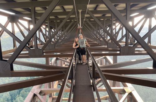 A couple stands on a metal bridge high above a valley, surrounded by steel beams and a scenic view.