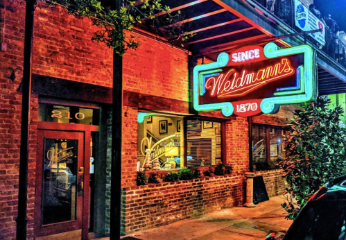 A vibrant neon sign for Weldmann's restaurant, established in 1870, glowing against a brick facade at night.