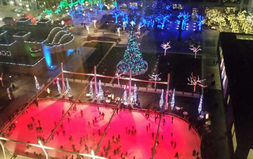 Aerial view of an ice skating rink surrounded by colorful holiday lights and a decorated Christmas tree.