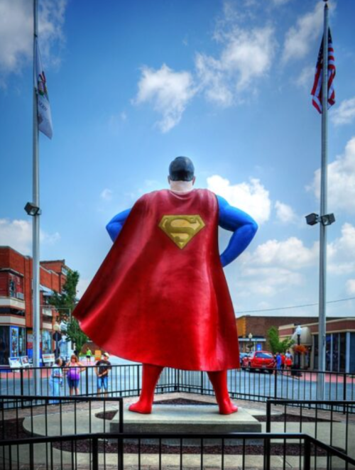 A large Superman statue stands with its back to the viewer, wearing a red cape, against a blue sky and flags.
