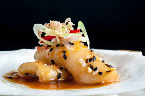 A beautifully plated dish featuring tender seafood drizzled with sauce, garnished with herbs and sesame seeds.