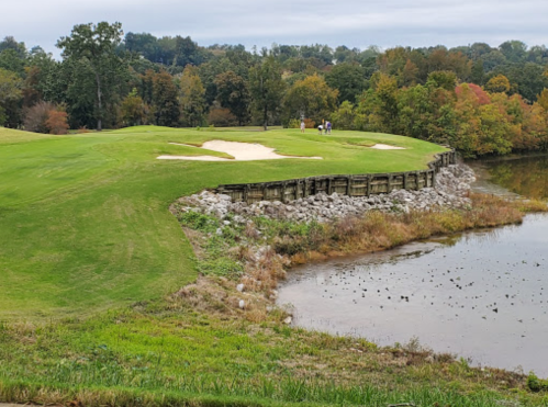 A scenic golf course with a sand trap, surrounded by trees and a calm water body, under a cloudy sky.
