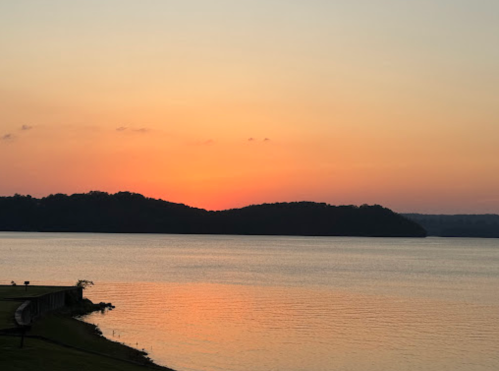 Sunset over a calm lake, with silhouettes of hills and a colorful sky reflecting on the water's surface.