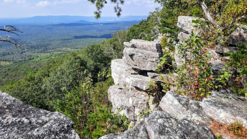 Rocky outcrop overlooking a lush green valley and distant mountains under a clear blue sky.