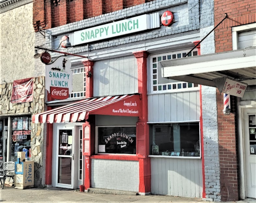 Exterior of Snappy Lunch, a colorful diner with a red and white awning, featuring a Coca-Cola sign and vintage decor.