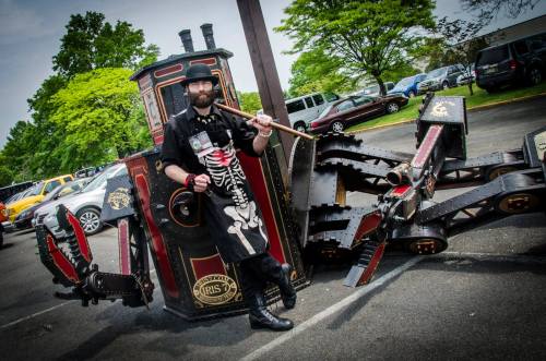 A man in a skeleton costume poses next to a creatively designed steam engine vehicle at an outdoor event.