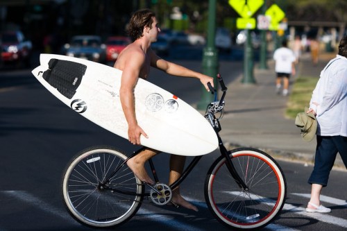 A shirtless man rides a bike while carrying a surfboard, with pedestrians and street signs in the background.
