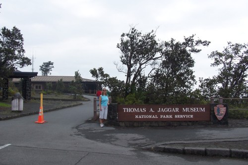 A person stands near the entrance sign of the Thomas A. Jaggar Museum, surrounded by trees and a cloudy sky.