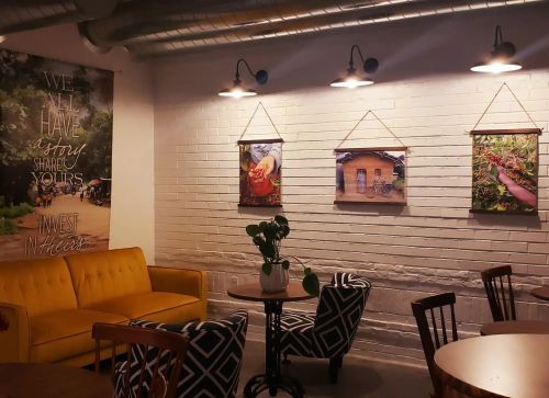 Cozy café interior with yellow couch, patterned chairs, and framed photos on a white brick wall. Inspirational wall art visible.