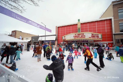 A lively winter festival scene with people ice skating in front of a theater, surrounded by snow and festive decorations.