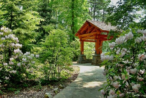 A stone pathway leads to a vibrant orange gazebo surrounded by lush greenery and blooming flowers.