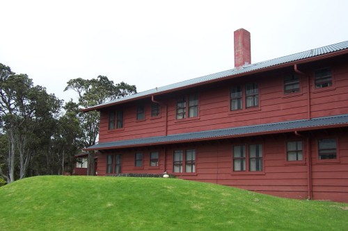 A red wooden house with multiple windows, set on a grassy hill surrounded by trees under a cloudy sky.