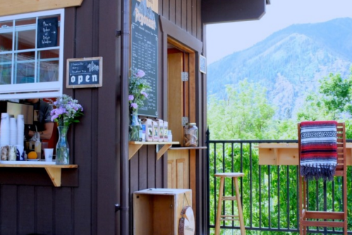 A cozy café with a mountain view, featuring an open sign, flowers, and a colorful blanket draped over a chair.