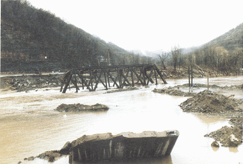 A dilapidated wooden bridge partially submerged in muddy water, surrounded by a hilly landscape.