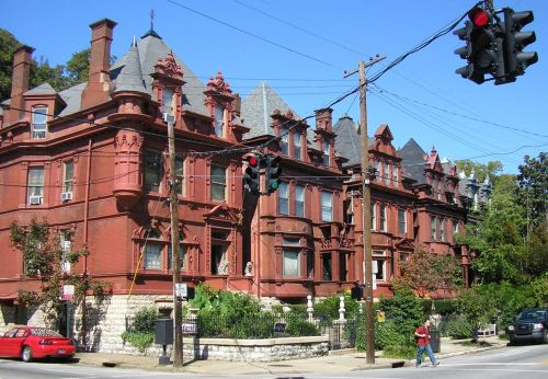 Historic red brick buildings with ornate architecture, surrounded by greenery, under a clear blue sky and traffic light.