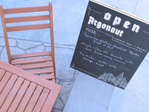 A chalkboard sign outside a café lists menu items, with a wooden chair beside it on a stone surface.