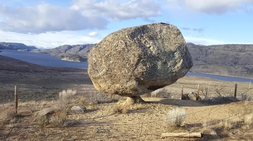 A large boulder balanced on a small base, surrounded by a scenic landscape with a lake and hills in the background.