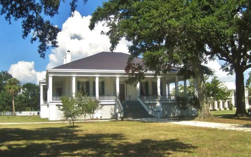 A large white plantation-style house with a dark roof, surrounded by trees and a grassy area under a blue sky.
