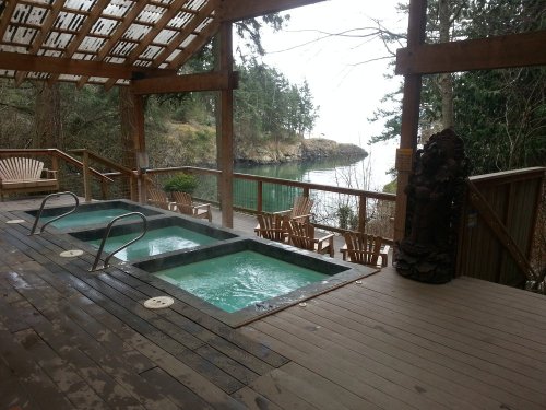 A serene outdoor spa area with two hot tubs overlooking a calm water view and lush greenery.