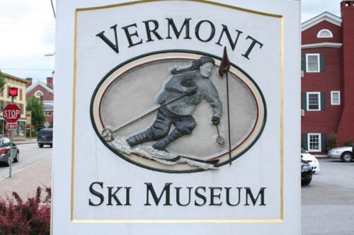 Sign for the Vermont Ski Museum featuring a skier in motion, with a red building in the background.