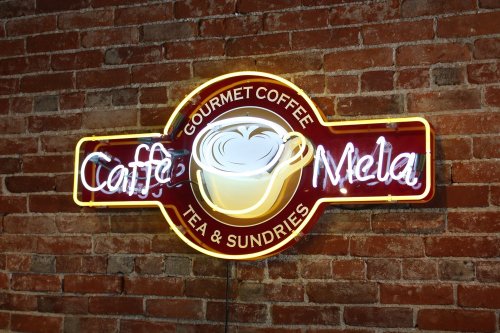 Neon sign for "Caffe Mela" featuring a coffee cup, set against a brick wall.
