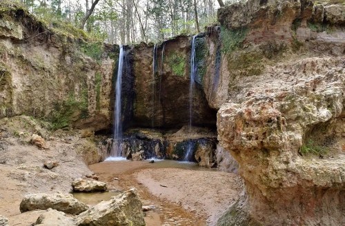 A serene waterfall cascades over rocky cliffs into a sandy area, surrounded by lush greenery and natural rock formations.