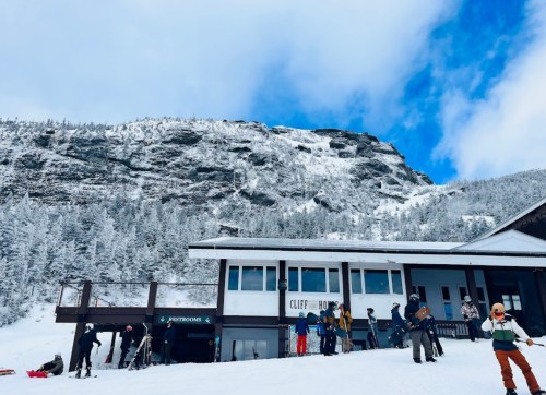 Skiers gather outside a lodge with snowy mountains in the background under a partly cloudy sky.