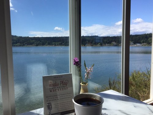 A cup of coffee sits on a table by a window, overlooking a serene lake and green hills, with flowers in a vase.