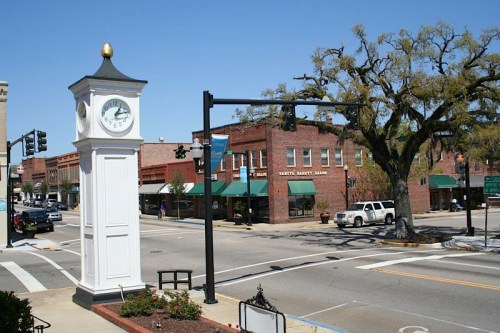 A clock tower stands at an intersection in a small town, with shops and trees in the background under a clear blue sky.