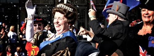 A joyful crowd celebrates, with a woman in a crown waving, surrounded by flags and confetti.