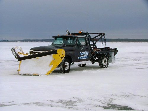 A black truck with a large yellow snow plow on a frozen lake, surrounded by snow and gray skies.