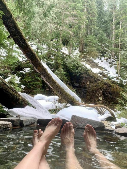 Two pairs of feet relax in a natural hot spring, surrounded by lush greenery and patches of snow.