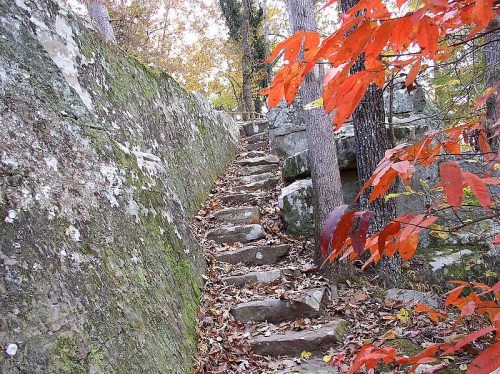 A rocky path leads upward through autumn foliage and trees, with vibrant red leaves in the foreground.