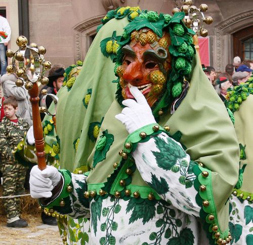 A person in a colorful mask and leafy costume holds a staff, surrounded by a festive crowd during a celebration.
