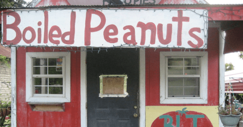 A small red building with a sign reading "Boiled Peanuts" and a door, decorated with lights and a tomato mural.