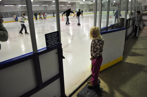 A young girl in a pink outfit stands by the rink, watching skaters glide on the ice in a busy skating rink.