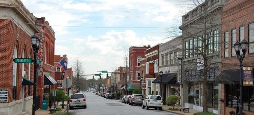 A charming small-town street lined with brick buildings, shops, and trees under a partly cloudy sky.