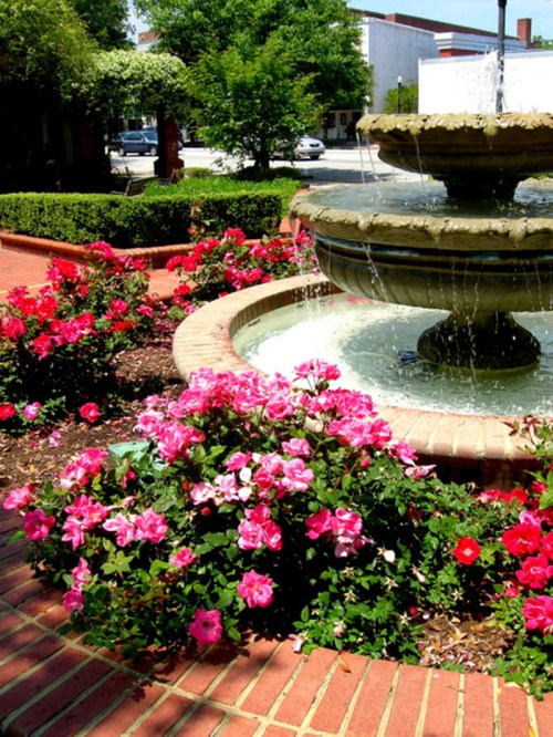 A decorative fountain surrounded by vibrant pink and red flowers in a sunny garden setting.