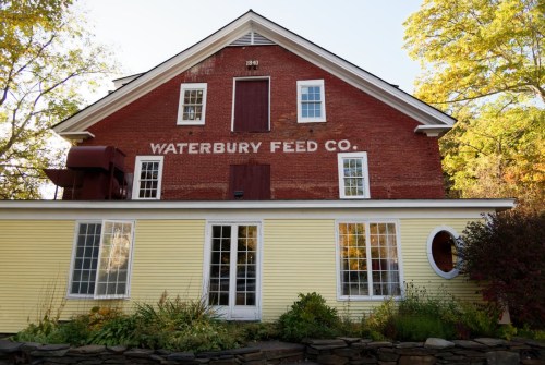 A historic red brick building with "WATERBURY FEED CO." painted on it, featuring large windows and a yellow addition.