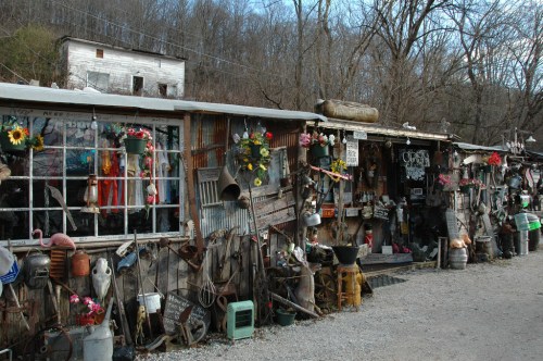 A rustic shop front adorned with flowers, signs, and various vintage items, set against a wooded backdrop.