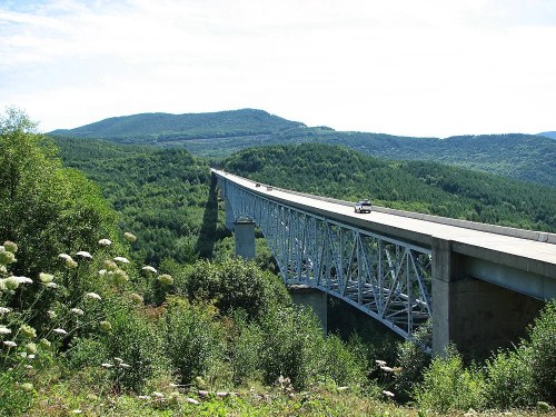 A long bridge spans a lush green valley, with mountains in the background and a car driving across it.