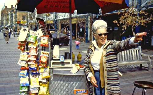 An elderly woman in sunglasses gestures beside a snack cart filled with chips, under a large umbrella on a busy street.