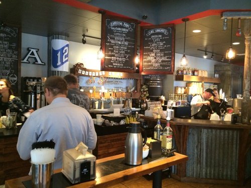A busy bar with patrons ordering drinks, featuring a chalkboard menu and rustic decor.