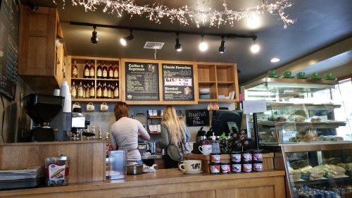 Two baristas work behind a wooden counter in a cozy café, with shelves of drinks and pastries visible.
