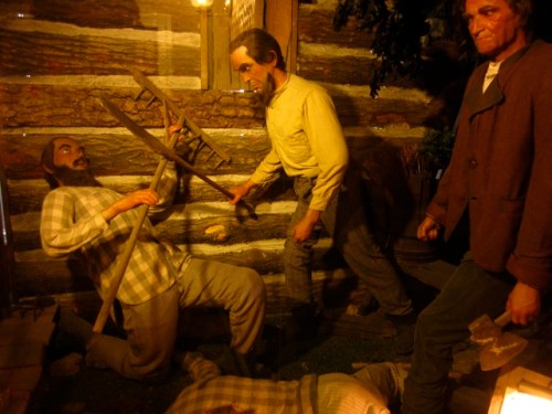 Wax figures depicting historical figures in a rustic setting, engaged in labor with tools and a log cabin backdrop.