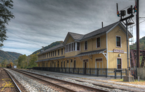 Historic yellow train station beside railroad tracks, set against a backdrop of mountains and a cloudy sky.