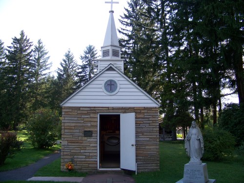 A small stone chapel with a cross on top, surrounded by trees and a statue of a figure nearby.