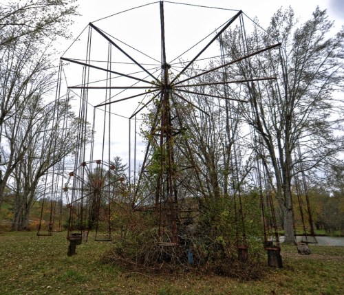 An old, rusted Ferris wheel overgrown with plants, surrounded by trees in a cloudy park setting.